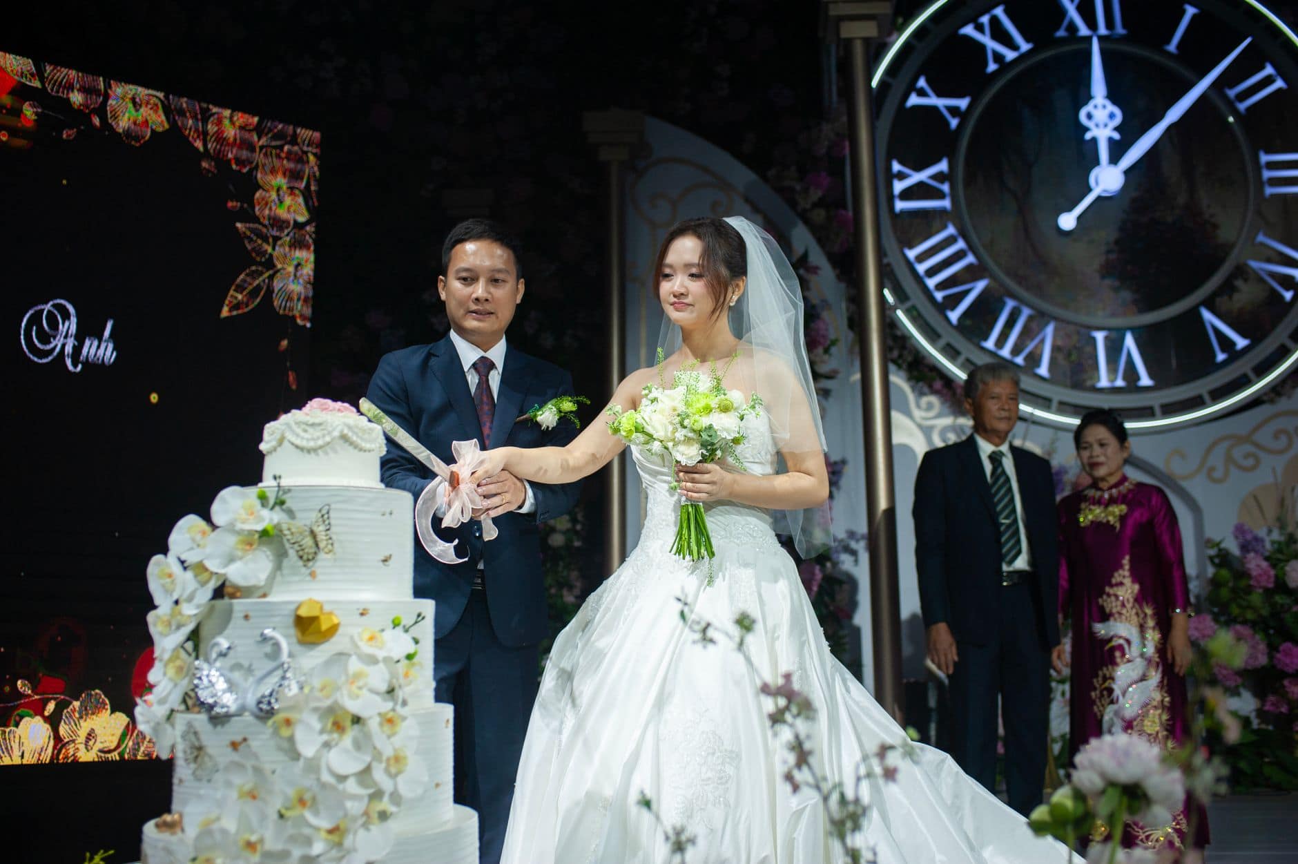Bride and groom cutting wedding cake with large clock backdrop in elegant setting.