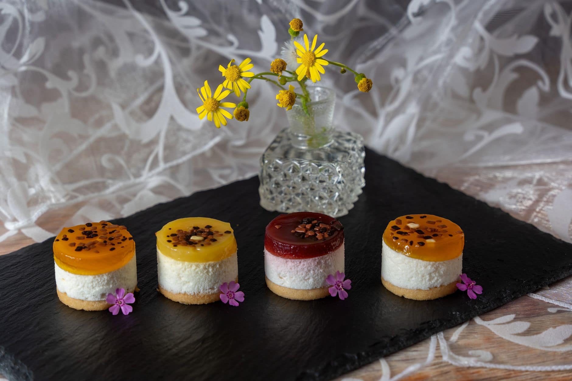 Assorted mini fruit cheesecakes with vibrant toppings presented on a slate board with flowers.