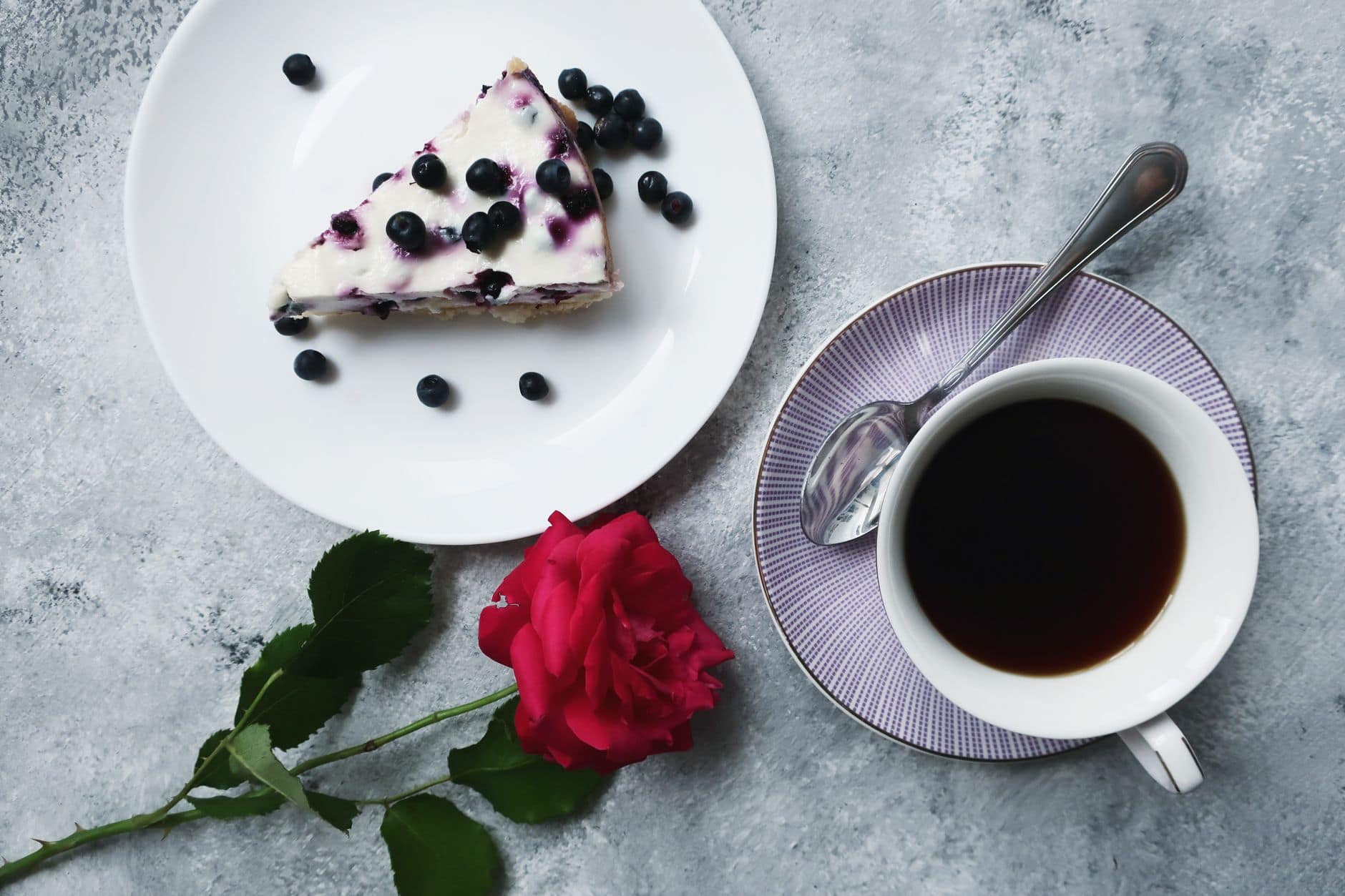 A delectable slice of blueberry cheesecake paired with black coffee and a red rose on a minimalist background.