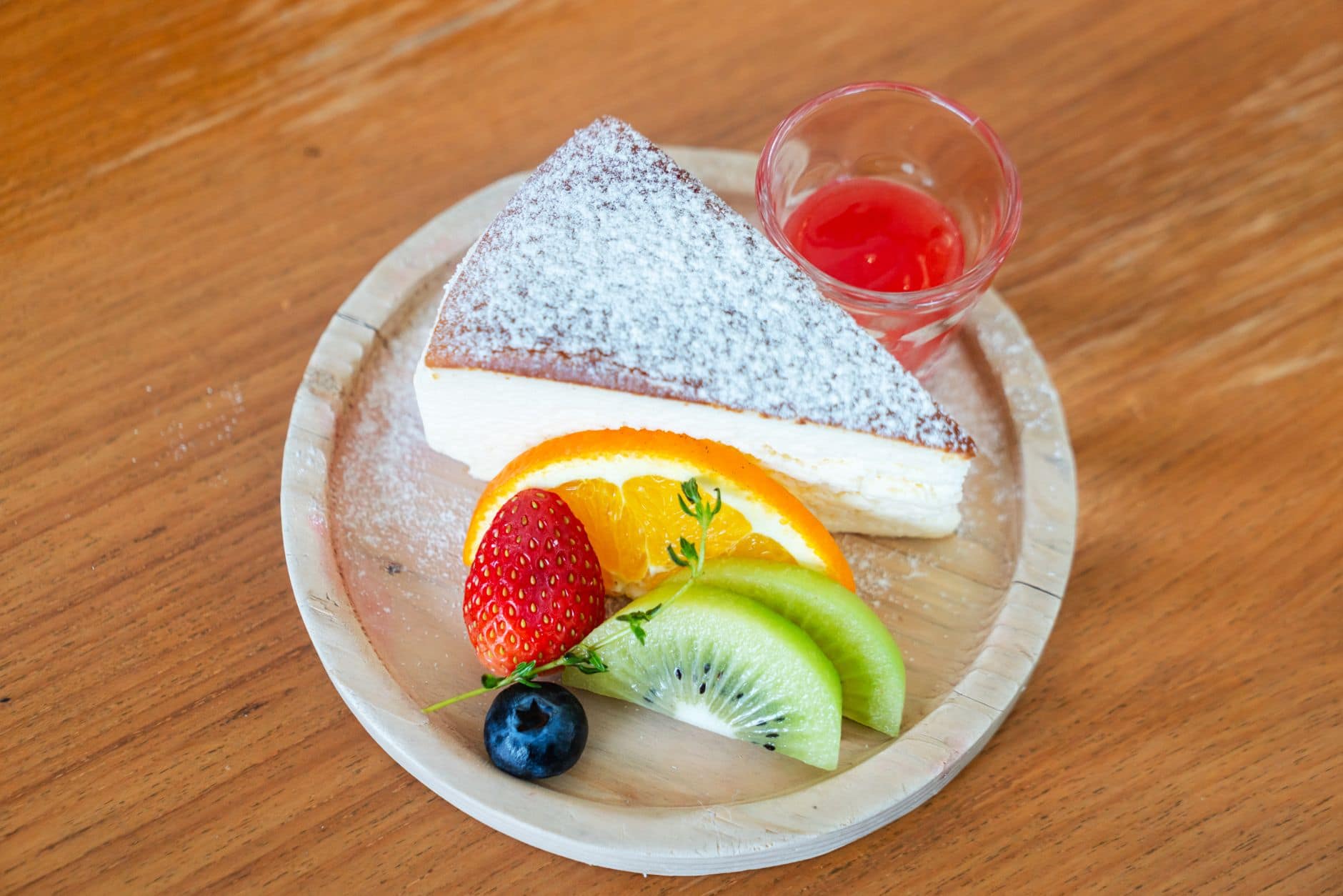 A slice of cheesecake with powdered sugar and fresh fruits on a wooden plate.