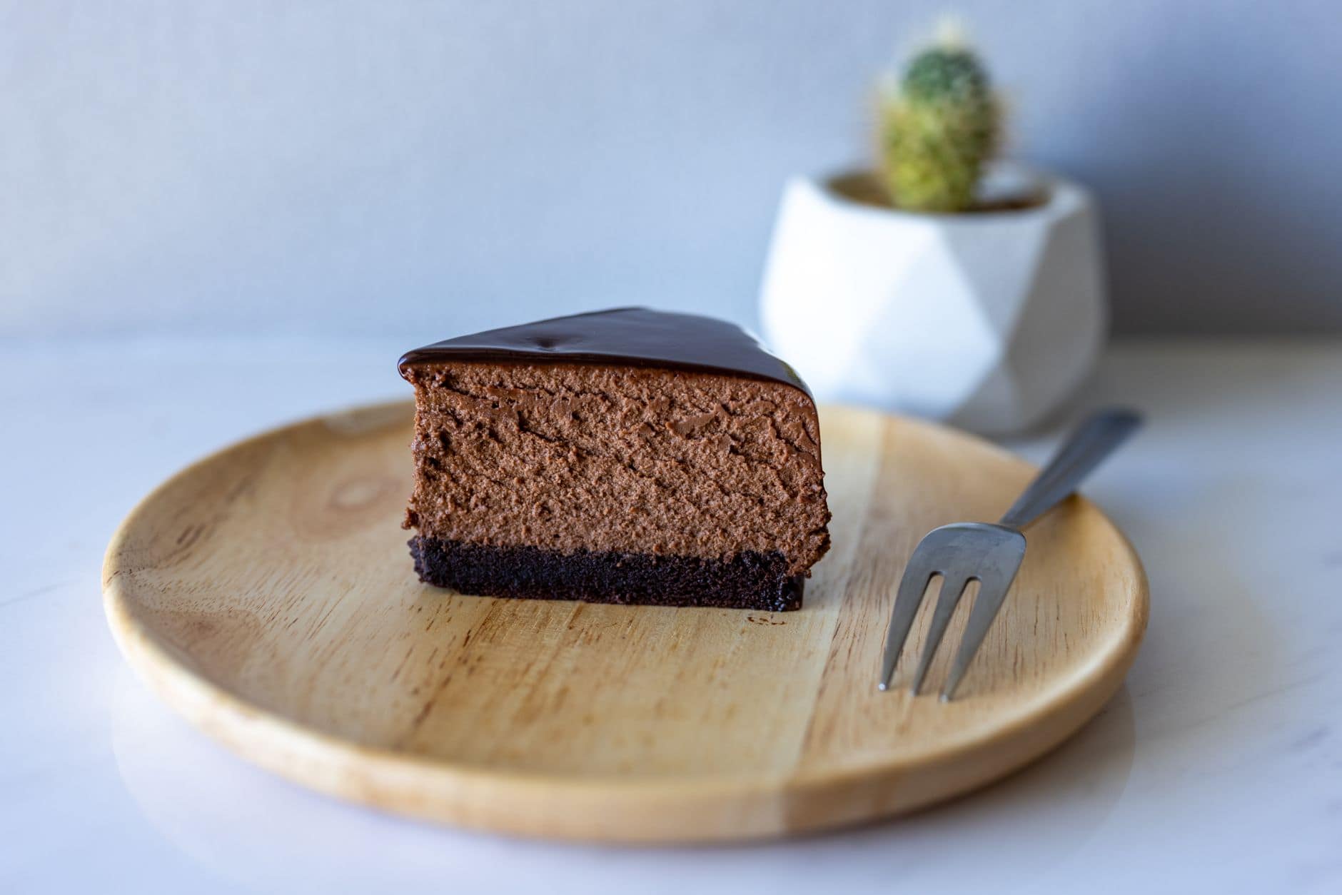 Indulgent slice of chocolate cheesecake on a wooden plate with a cactus in the background.